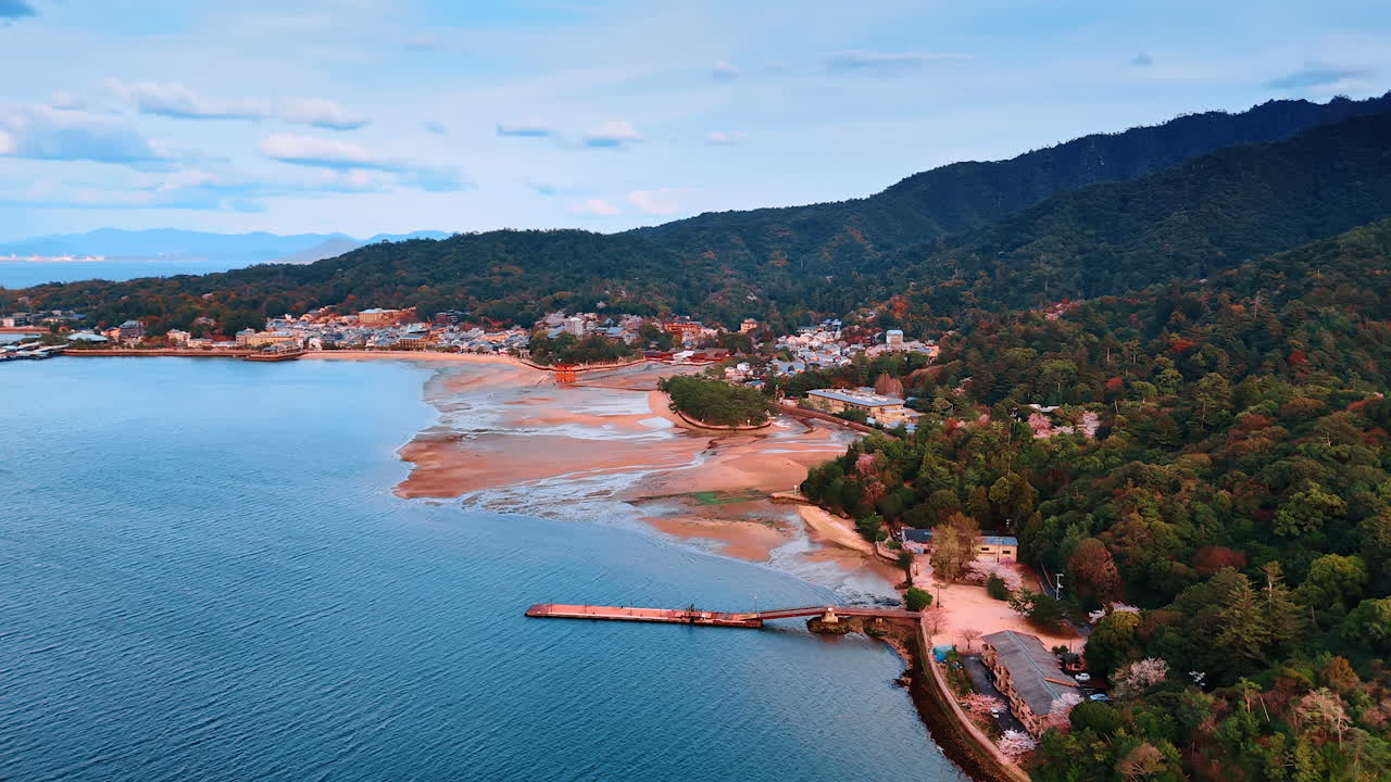 Period of low tide at the shore with famous red Grand Torii Gate at the Miyajima island, Japan. Drone footage approaching landmark revealing view on the inhabited area and gorgeous wooded mountains.