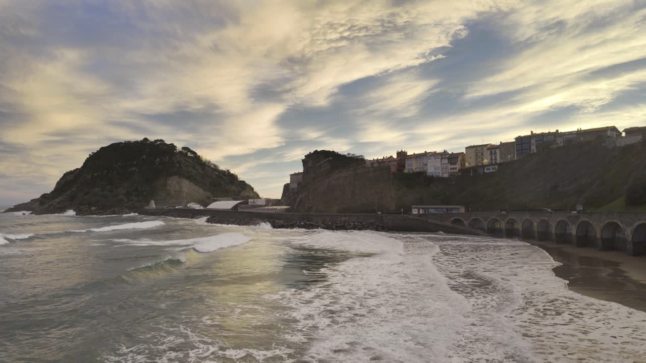 Bellissimo paesaggio costiero di Getaria nei Paesi Baschi, Spagna, con le onde dell'oceano che si infrangono sulla spiaggia sabbiosa sotto un cielo nuvoloso e drammatico al tramonto, mostrando l'iconica montagna di San Anton.