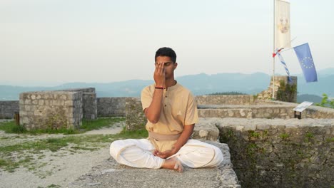 slow-motion of young indian male doing nadhi shuddhi meditation at sunrise on stone wall