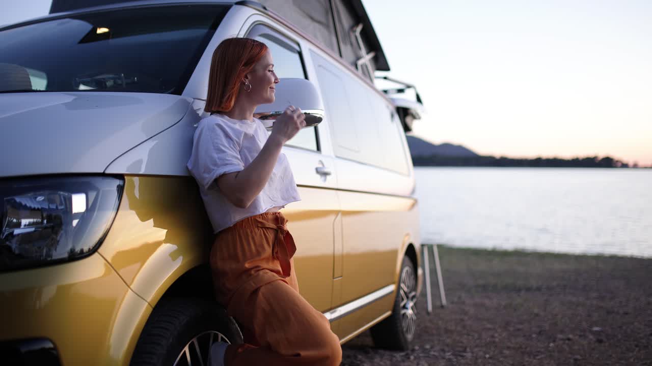 Giovane donna appoggiata a un camper in riva al lago al tramonto