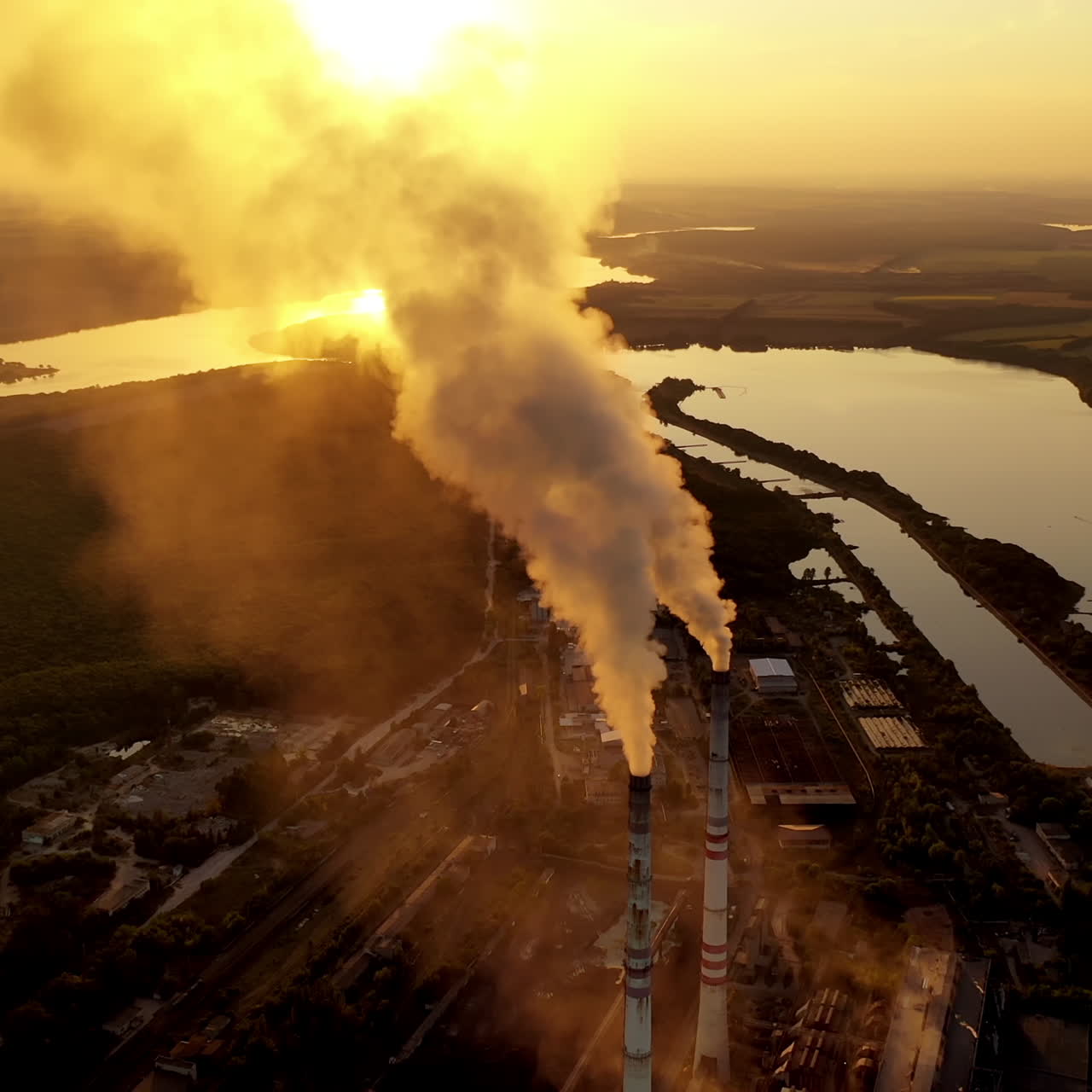 Industrial plant near the river. Metallurgical factory with large pipes. Dirty emissions coming out of pipes into the air at sunset. Aerial view.