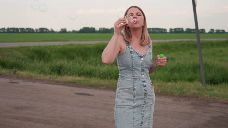full body young woman in denim dress standing on rural dirt road holding bubble bottle in one hand blowing bubbles into warm evening air over open green field with distant trees