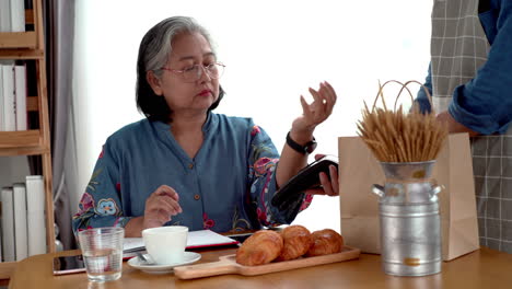 Active-senior-Asian-woman-working-in-a-coffeeshop-using-a-mobile-device,-smartwatch-for-NFC,-QR-code-scanning,-contactless-payment-to-pay-for-coffee-and-snack