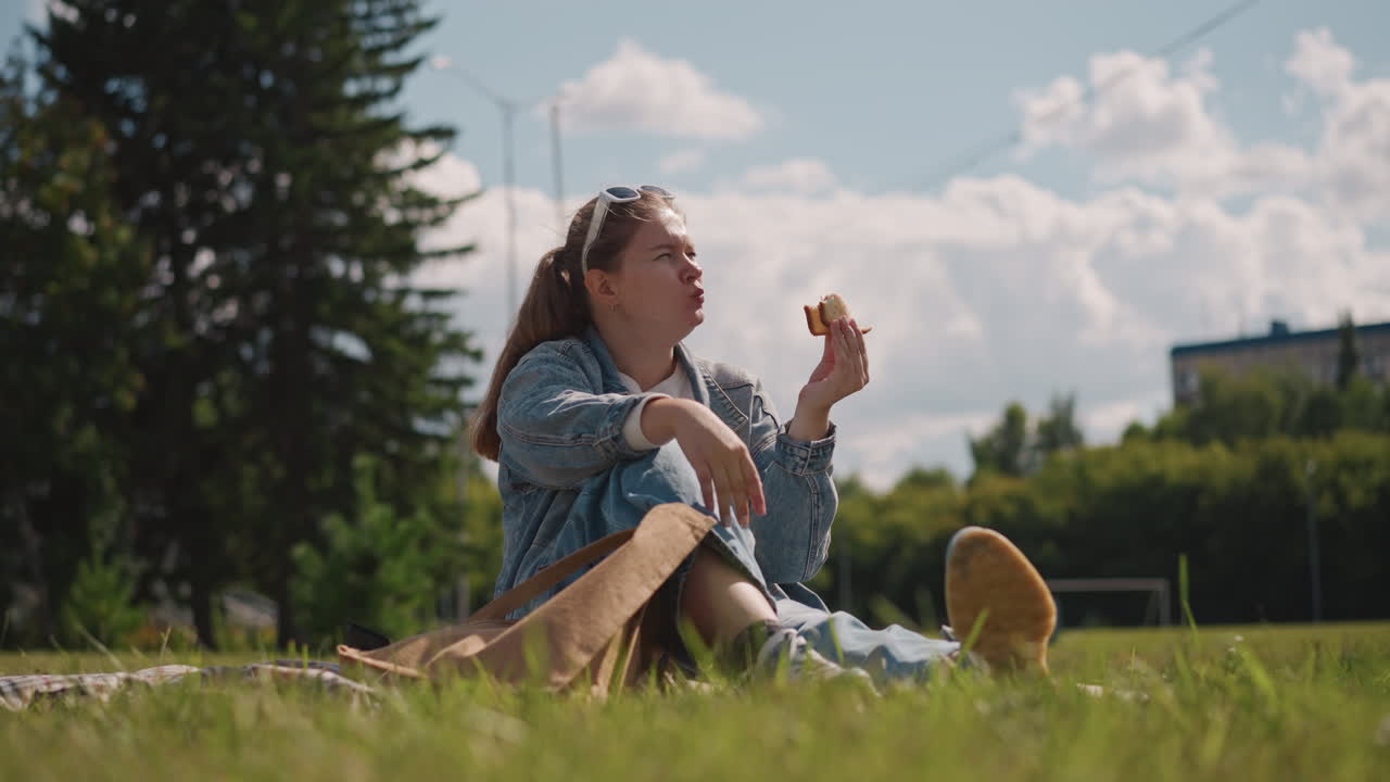 Mujer sentada en un parque de césped disfrutando de un cruasán, tarde soleada, ajustándose las gafas de sol, postura relajada, chaqueta de mezclilla casual, bolso de tela cerca, edificios distantes y árboles altos, nubes suaves.