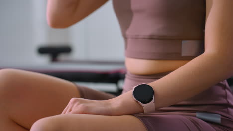vista parcial de la parte inferior del cuerpo de una mujer en traje de gimnasio usando un reloj inteligente mientras bebe agua, momento relajado después del entrenamiento con fondo borroso con varios equipos de gimnasia