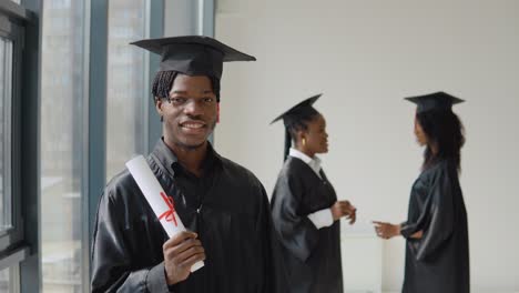 un joven afroamericano está de pie en primer plano y mira a la cámara con un diploma en las manos. dos mujeres afroamericanas se comunican entre sí en el fondo.