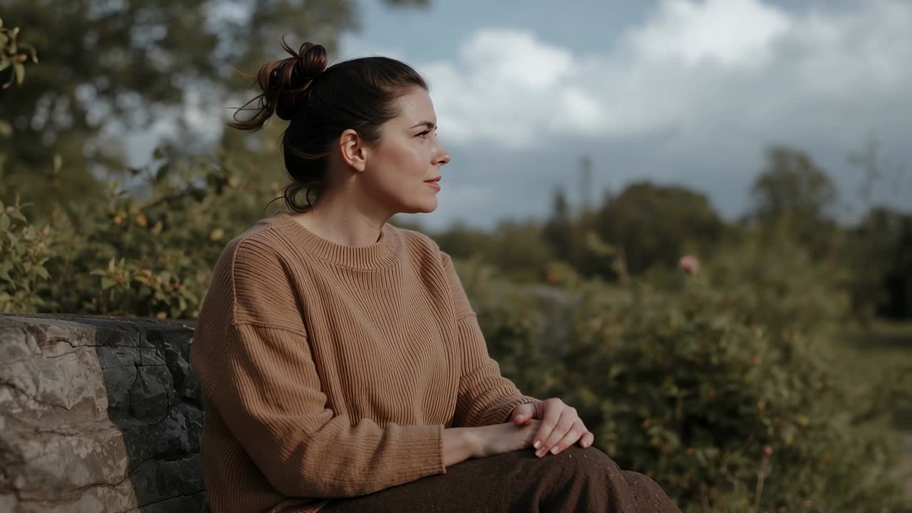 Mulher sentada com as mãos juntas em um banco de pedra, sorrindo e virando a cabeça, contemplando as árvores com um suéter bege.