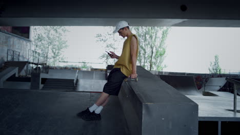 un adolescente relajado con un teléfono inteligente sentado en una tabla de patinaje en un parque de patinaje.