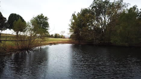 aerial drone backward moving shot over small lake along green grasslands and trees on a cloudy day