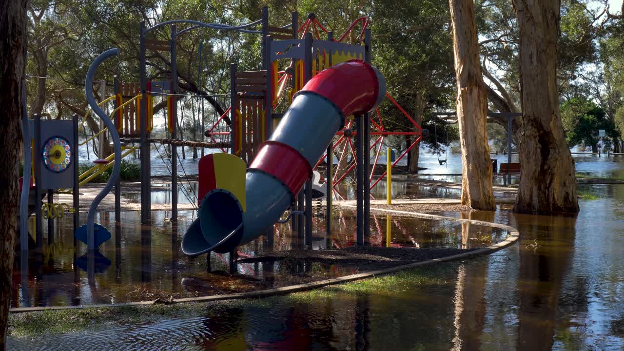 Slow motion landscape of children's playground equipment with slide covered in flood water river system lake overflow and trees in Budgewoi Central Coast Australia risk hazard damage weather disaster
