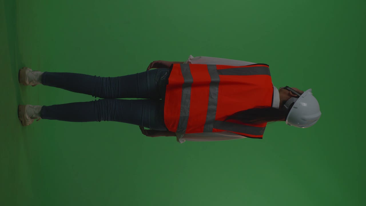 동양 여성 엔지니어가 안전 헬을 착용해 녹색 스크린 배경 스튜디오에 서 있는 동안 주변을 둘러보고 있는 모습.