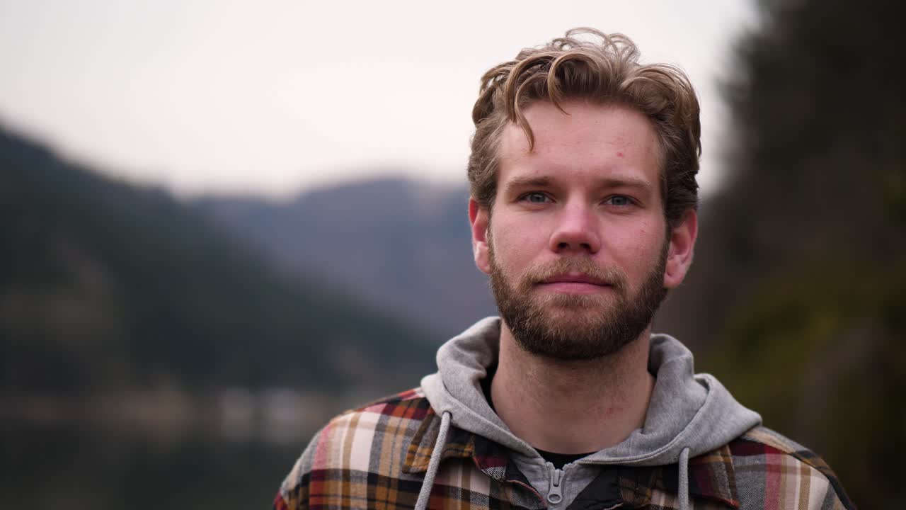 A young bearded man with wavy hair filmed in slow motion walks between hills and forest along a small path