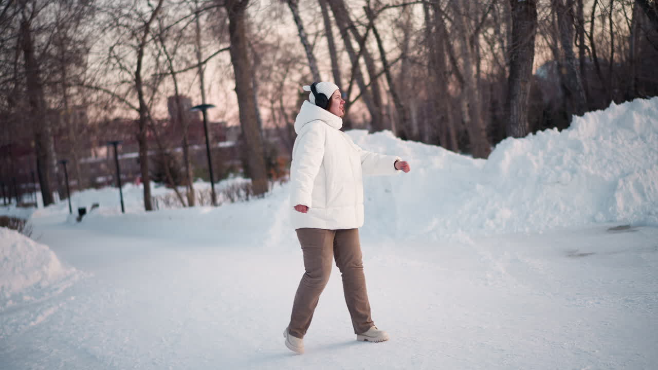 Girl wearing tinted goggles, white puffer coat and beanie dances and sings cheerfully with headset under sunset light in snowy park surrounded by bare trees capturing joyful winter movement