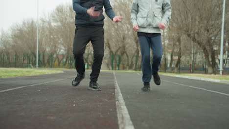 a coach guides a young boy through jogging exercises on a stadium track, by instructing him to lift his legs higher with bare trees and bar fence in the background