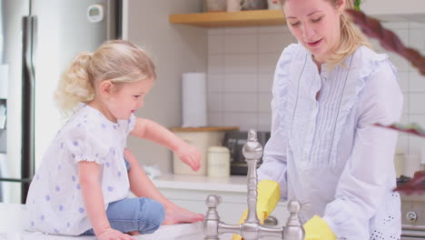 mother wearing rubber gloves at home in kitchen with young daughter having fun as they do washing up at sink- shot in slow motion