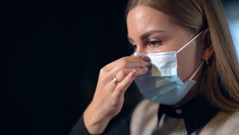 young woman in mask sitting half-turned to the camera. long-haidred lady touching her mask at nose-bridge. close up.