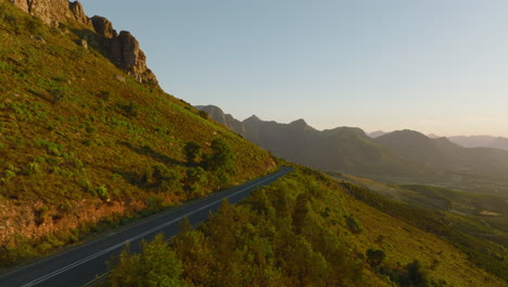 Adelante-Vuela-A-Lo-Largo-De-La-Carretera-Que-Conduce-A-Través-Del-Paisaje-Montañoso.-Escena-Iluminada-Por-La-Luz-Brillante-Del-Sol-Poniente.-Sudáfrica