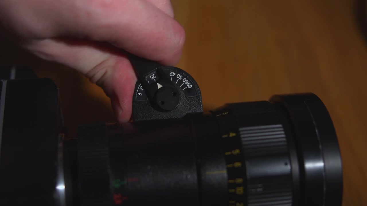 Nahaufnahme einer Hand, die den Einstellring an einem Kameraobjektiv dreht, mit detaillierten Markierungen, Metall- und Kunststoffoberflächen und einem warmen, unscharfen Innenhintergrund.