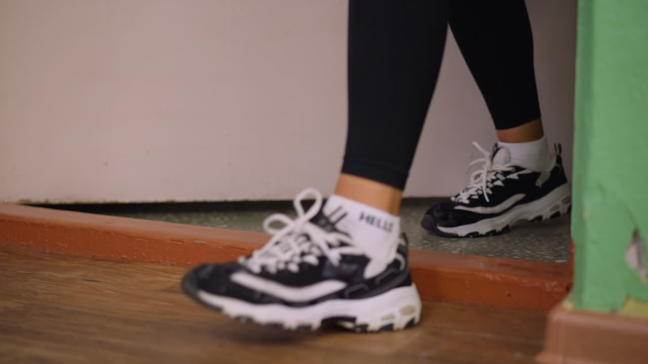 Closeup of leg in sneakers stepping out through doorway, casual movement emphasized with focus on shoe, floor, and threshold details, indoor setting captured with soft background highlighting action