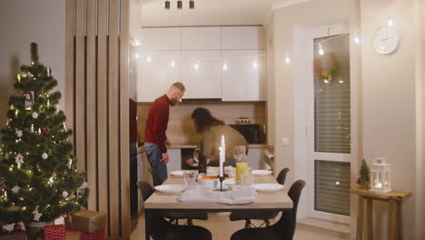 brunette woman taking turkey out of the oven, red haired man closes the oven and the children come to the table and the whole family sits for christmas family dinner