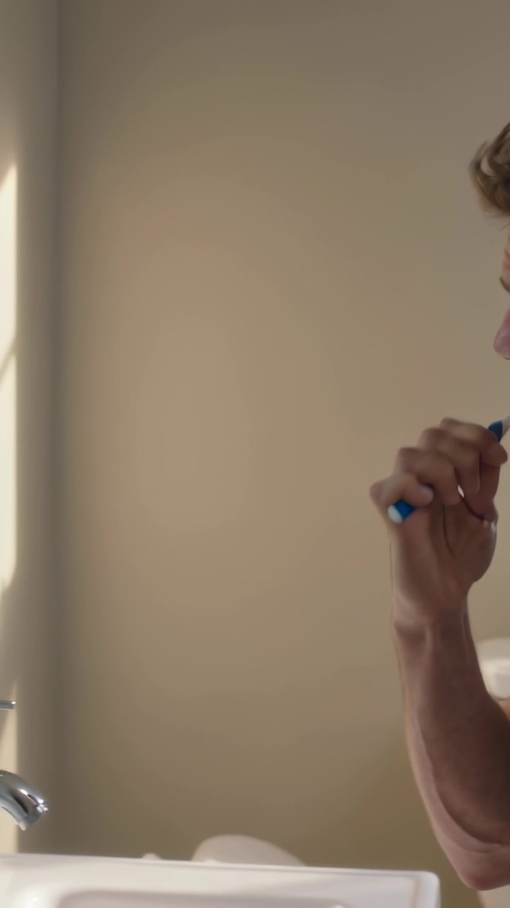 Vertical video: Panning to man grey tee brushing teeth at sink with blue brush for care, copy space