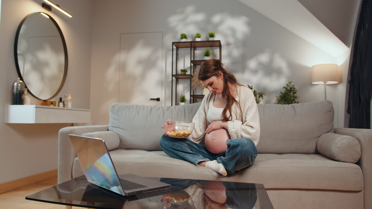 Gravid kvinde sidder på sofaen med en skål popcorn og ser på en bærbar computer med et kedsommeligt udtryk gravid