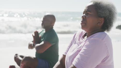 happy senior african american couple doing yoga and meditating at beach, in slow motion