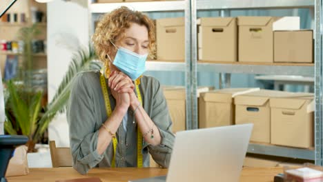 young caucasian designer woman in facial mask sitting at desk and videochatting on laptop in clothing shop warehouse