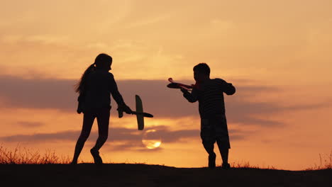 Silhouettes-Of-A-Girl-And-A-Boy-Playing-Together-With-Airplanes-At-Sunset-A-Happy-And-Carefree-Child