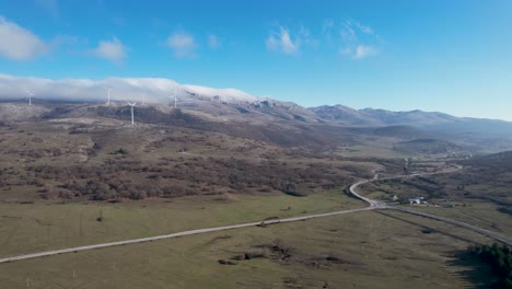 Wunderschöne-Luftaufnahme-Der-Kroatischen-Landschaft-Mit-Windturbinen,-Die-Im-Hintergrund-Erneuerbare-Energie-Erzeugen,-Und-Einer-Leeren-Straße-In-Der-Region-Lika-In-Kroatien,-Europa