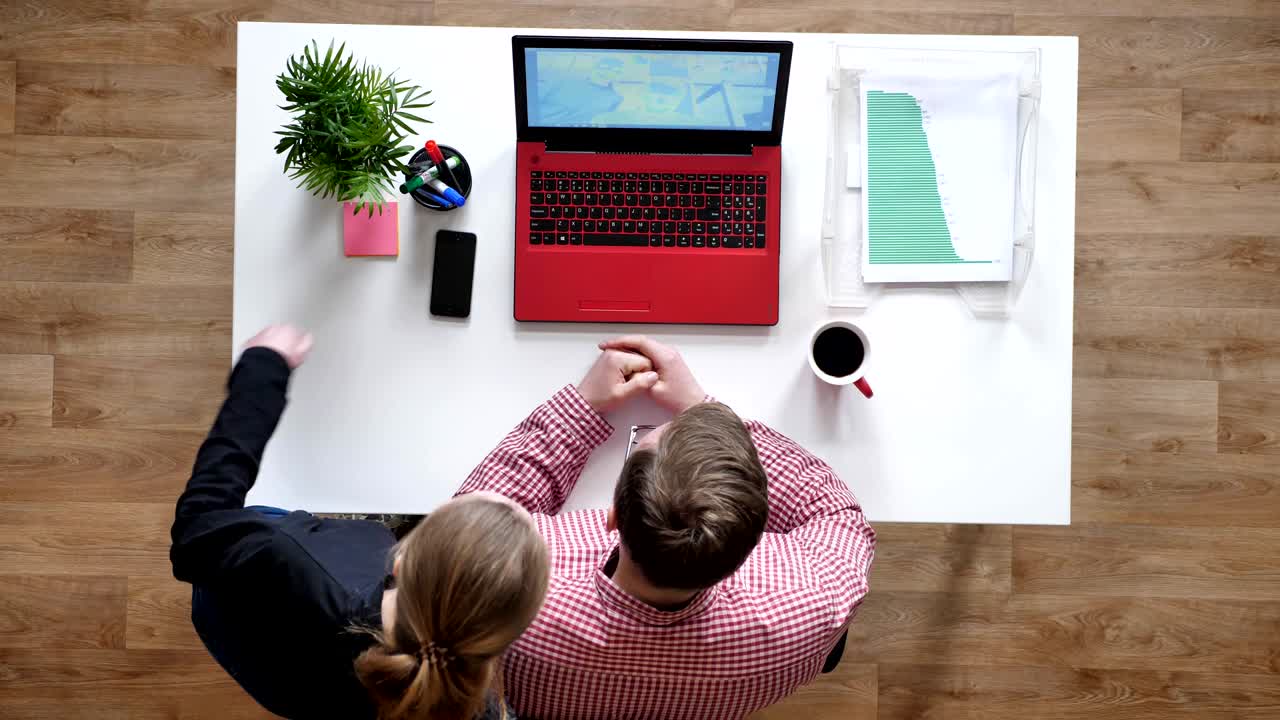 joven con gafas invitando a una mujer a hablar con alguien a través de una computadora portátil, topshot, escritorio con café y documentos
