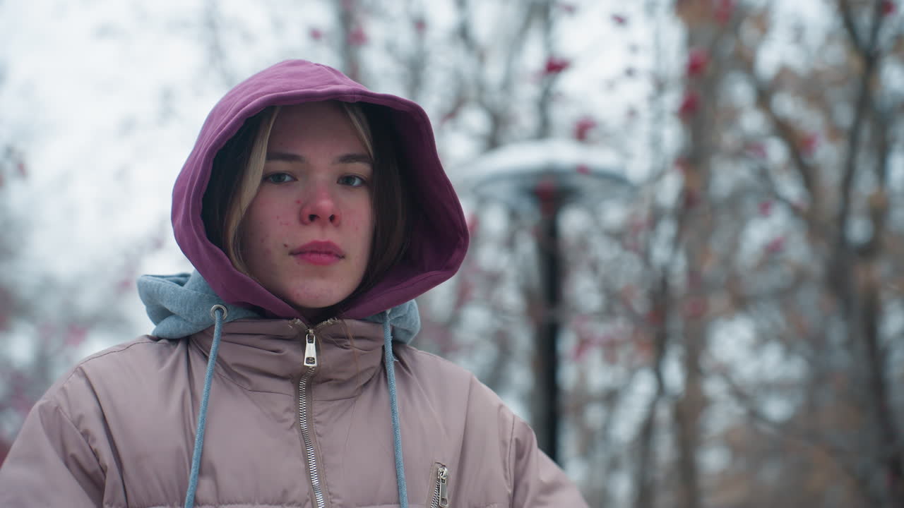 젊은 여성이 자신감있는 포즈로 후디를 조정하고 카메라를 바라보고, 나무와 가을 잎, 패션 및 겨울 진동으로 흐릿한 배경