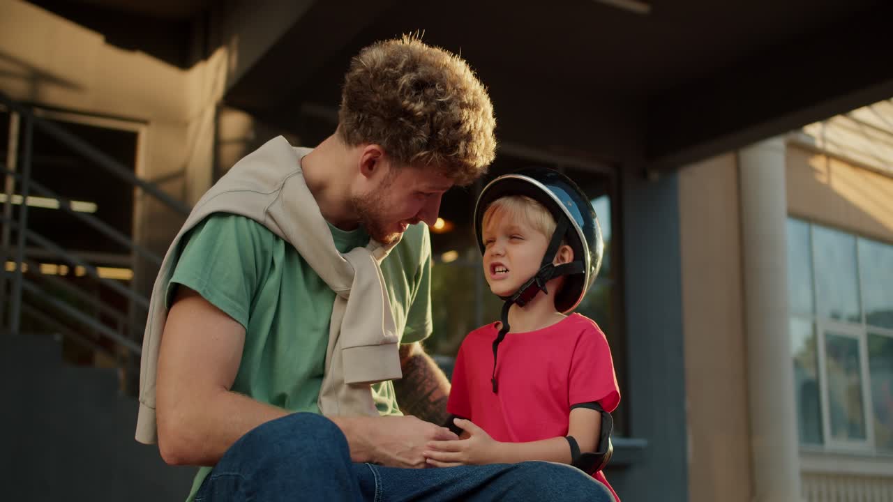 padre feliz con el cabello rizado en una camiseta verde se sienta cerca de su hijo pequeño presa en un casco negro y en una camisa roja durante el tiempo soleado por la noche en verano