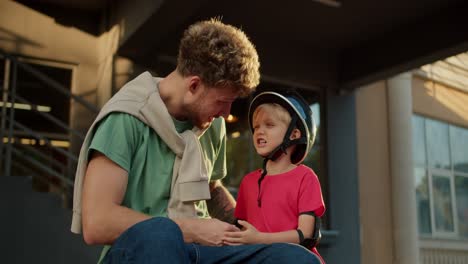 Happy-father-with-curly-hair-in-a-green-t-shirt-sits-near-his-little-son-dam-in-a-black-helmet-and-in-a-red-t-shirt-during-sunny-weather-in-the-evening-in-summer