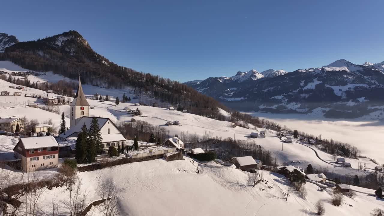 Village church with alpine homes on rolling white hills overlooking fog covering Walensee in Switzerland