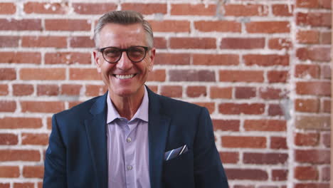 Portrait-Of-Smiling-Mature-Businessman-Standing-Against-Brick-Wall-In-Modern-Office