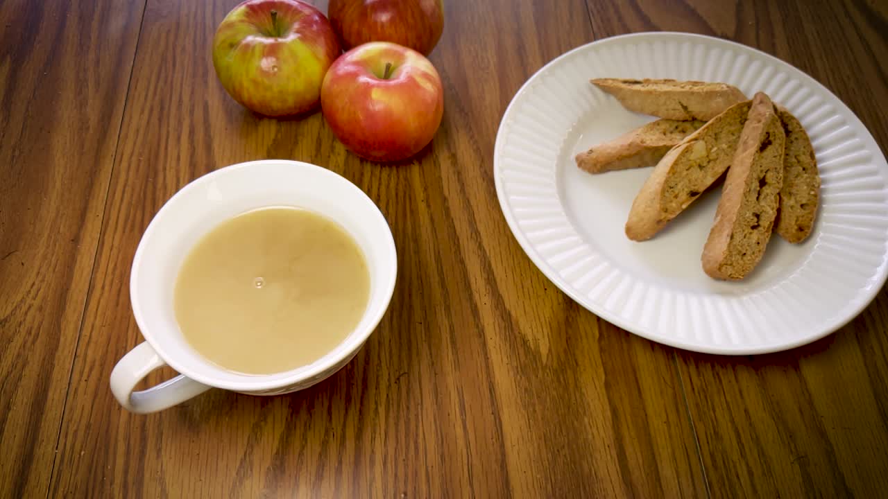 Slow motion shot of milk being poured into a cup of tea with apples and cookies in the background