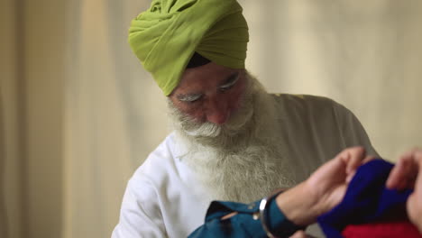 Fotografía-De-Estudio-De-Un-Hombre-Sikh-Mayor-Que-Ayuda-A-Un-Hombre-Sikh-Más-Joven-A-Atar-La-Tela-Para-Un-Turbante-Contra-Un-Fondo-Liso,-Filmada-En-Tiempo-Real-6