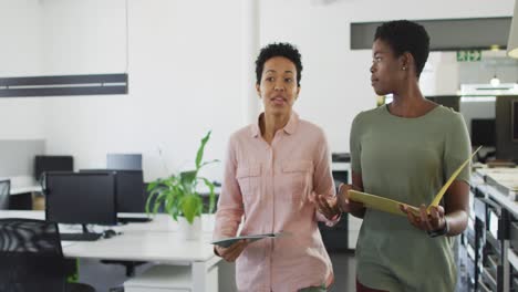 Two-diverse-businesswomen-walking-in-office-with-documents-and-talking