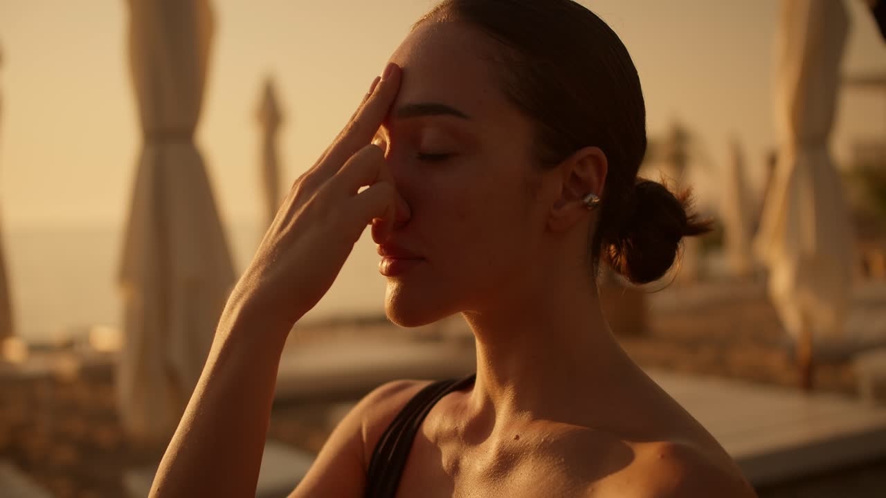 Close-up shot of a brunette girl meditating and putting her fingers on her forehead, Breath of Fire on a sunny beach during