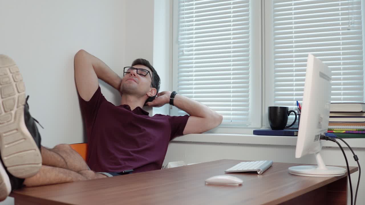 Young office worker in glasses relaxing in office with his legs on the table in the modern office. Break during the working time. Shot in 4k