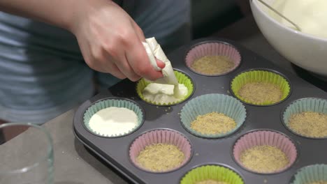 A-woman-using-a-piping-bag-filled-with-cheesecake-cream-filling-to-fill-muffin-holders,-which-sit-in-a-cupcake-pan,-to-make-cheesecake-cups