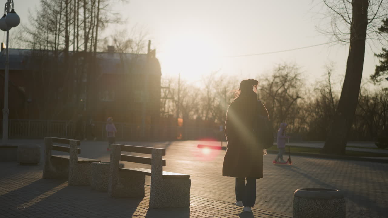 un retrato de una mujer con un abrigo marrón, vaqueros y zapatos blancos, llevando una mochila mientras camina por un parque durante la puesta de sol. la mujer parece infeliz, mientras los niños juegan a su alrededor