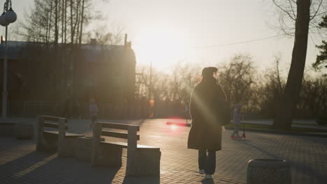 a backshot of a woman wearing a brown coat, jeans, and white shoes, carrying a backpack as she walks through a park during sunset. the woman appears unhappy, while children play around her