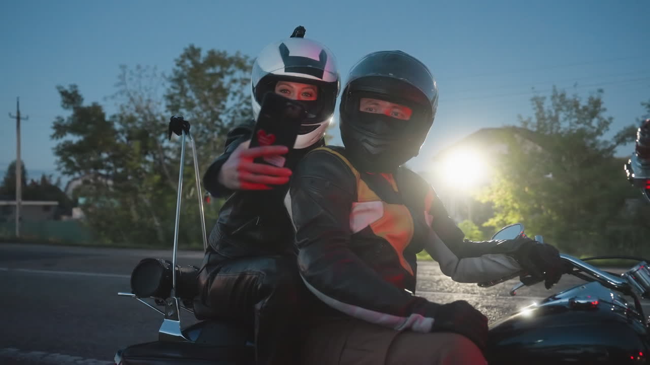 Couple wearing helmets and leather jackets sit on motorcycle at roadside during evening, woman extends arm holding phone for selfie while man looks forward