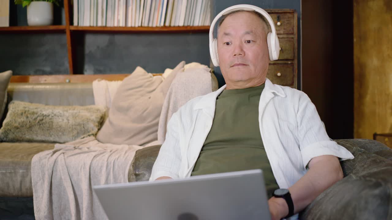 白いヘッドホンをつけ、ノートパソコンの再生画面を見ているアジア系のシニア男性がソファに座っている。