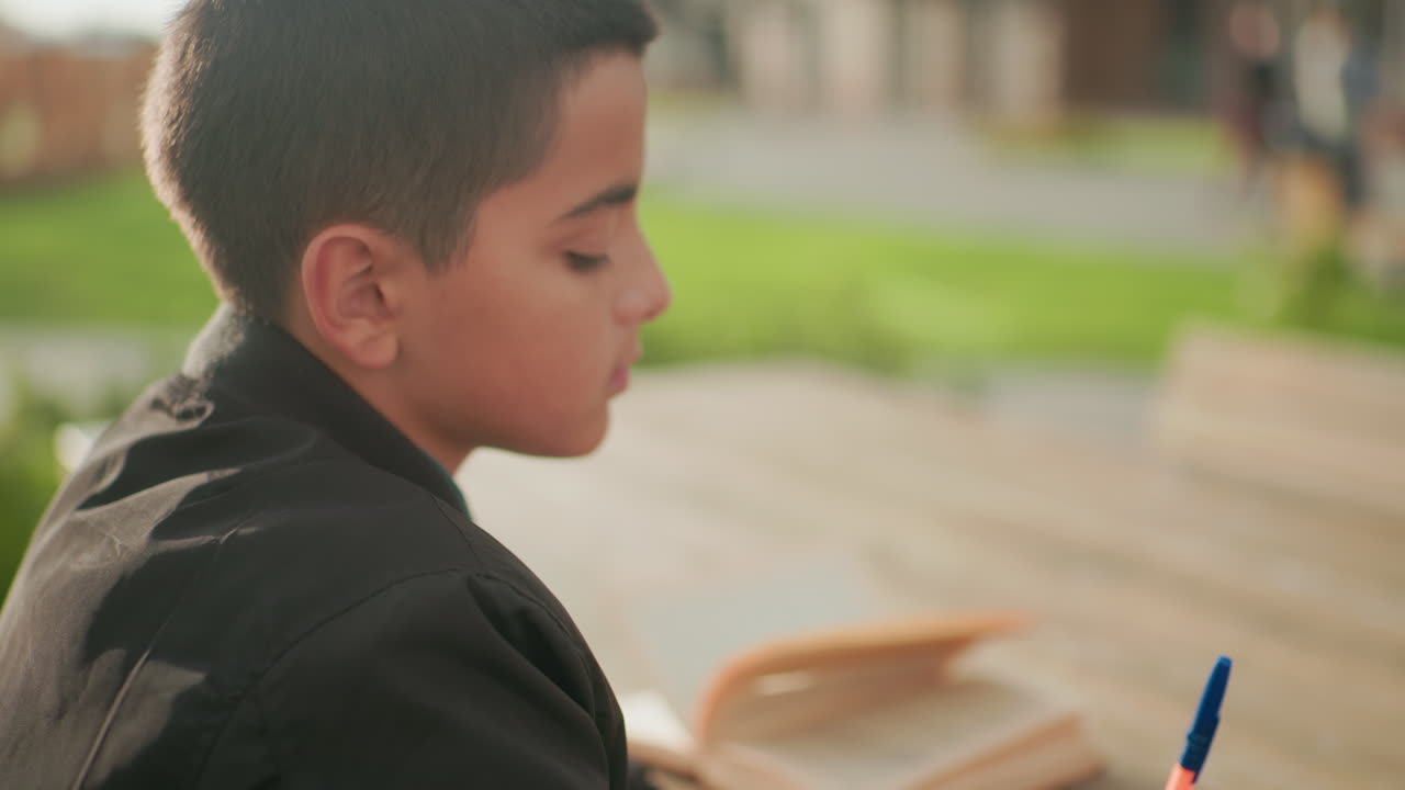 Close up side view of boy sitting outdoors at wooden table with open book and pen resting nearby, thoughtful expression as sunlight highlights background