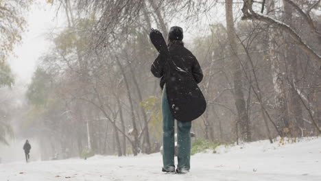 persona que lleva guitarra camina por un camino nevado, otra figura borrosa en la distancia, en medio de árboles de invierno y nieve que cae, capturando una atmósfera serena de invierno