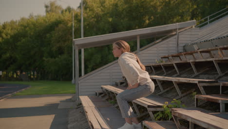 woman casually walks up rustic stadium seating on first row, wearing jeans and hoodie with relaxed posture, resting her head on hand, background includes empty bleachers, electric poles