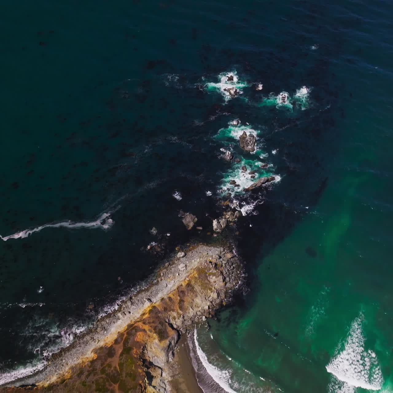 Flying over the rocky shore and moving to the azure waters of ocean. Little white waves splashing by the mountainous coastline. Top view